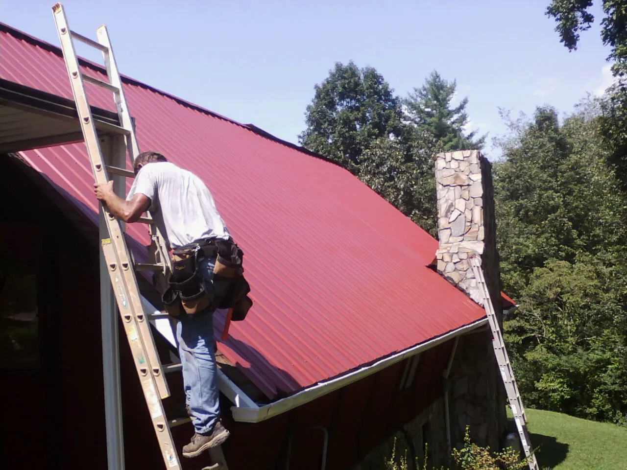 Roofer on a ladder beside a red metal roof and stone chimney — Soffit & Fascia Repair by Roofing Brothers in Northampton
